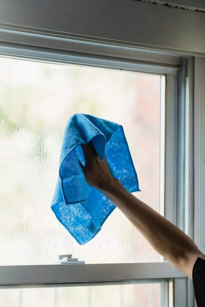A hand using a blue towel to clean a window indoors, showcasing housework.